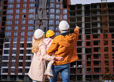Back view of man pointing at apartment building under constructing while standing next to wife and daughter on the street. Happy family looking at new home at construction site.の写真素材