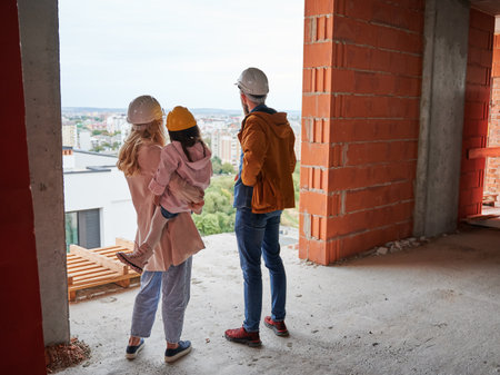 Back view of parents with daughter standing inside apartment building under construction. Man and woman with child wearing building helmets while enjoying city view from future home.の写真素材