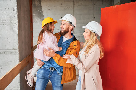 Cheerful man holding daughter and smiling while standing next to wife at construction site. Happy parents with child posing in apartment building under construction. New home purchase concept.の写真素材