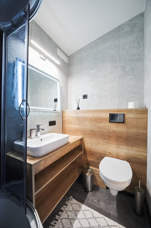 Clean, minimalist grey bathroom in slate with ceramic sink on wooden countertop and hanging toilet inside. Side view of basin and mirror illuminated with warm light in bathroom. Concept of interior.の写真素材