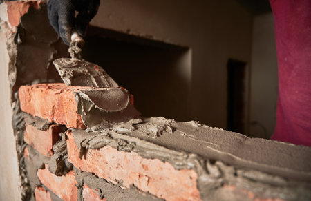 Close up of man hand in work glove laying brickwork in building under construction. Male worker applying cement mortar on bricks with trowel tool. Masonry construction concept.の写真素材