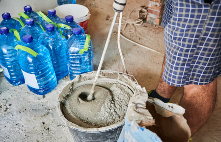 Close up of man builder in work gloves preparing sand-cement mix for bricklaying in building under construction. Male worker mixing mortar with electric cement concrete mixer.の写真素材