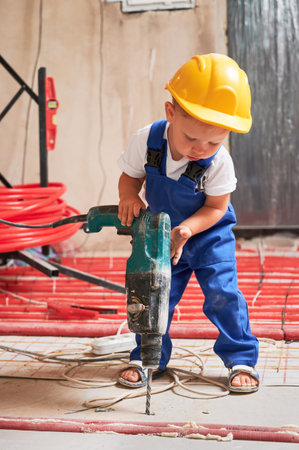 Child in safety helmet drilling floor with electric hammer drill at home during renovation. Kid construction worker using drilling machine while installing underfloor heating system in apartment.の写真素材