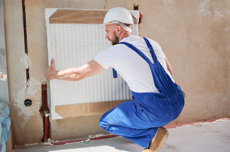 Male technician mounting heating radiator on the wall. Man wearing safety construction helmet and work overalls while working on home renovation.の写真素材