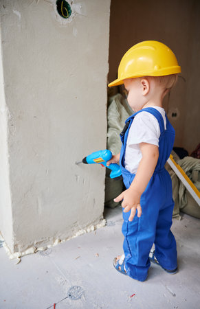Child drilling wall with toy electric drill tool in apartment under renovation. Kid in work overalls using drilling construction instrument at home during refurbishment.の写真素材