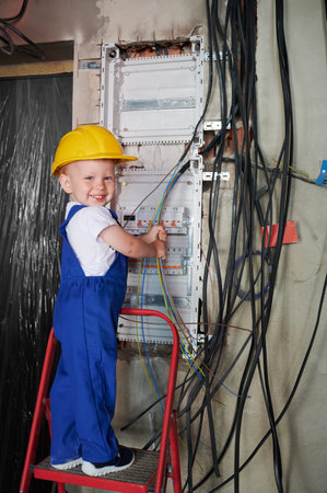 Smiling little boy in construction safety helmet holding electrical wires while fixing electric control panel in apartment. Kid electrician standing on ladder near electrical switchboard.の写真素材