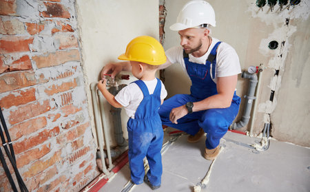 Father and little boy installing plumbing pipe system in apartment during renovation. Male plumber and kid in safety construction helmet checking pipe tap while working on home renovation.の写真素材