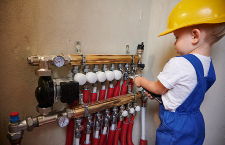 Baby boy in safety construction helmet using wrench tool while fixing plumbing installation with pipes and thermometer gauge. Kid plumber repairing plumbing pipe system in apartment under renovation.の写真素材