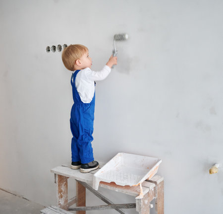 Little boy construction worker painting wall in apartment under renovation. Adorable kid in work overalls using paint roller while playing at future home.の写真素材