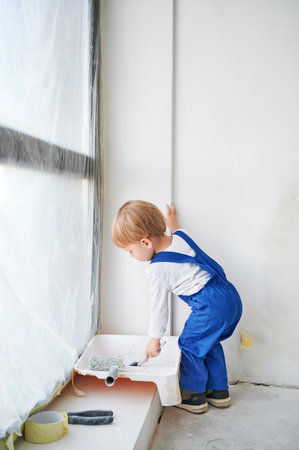 Little boy construction worker standing by the window and painting white wall in apartment. Kid in work overalls using paint brush while playing at home under renovation.の写真素材