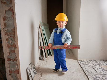 Full length of child construction worker holding spirit level instrument and looking at camera. Kid in safety helmet and work overalls standing by the wall in apartment under renovation.の写真素材