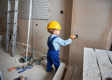 Full length of child construction worker painting wall with brush in apartment under renovation. Kid in safety helmet and work overalls playing at home, standing in room with tools and ladder.の写真素材