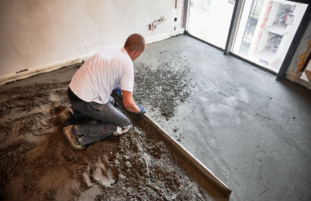 Male construction worker using screed rail while screeding floor in living room. Man flattening and smoothing surface with straight edge in apartment during renovation.の写真素材