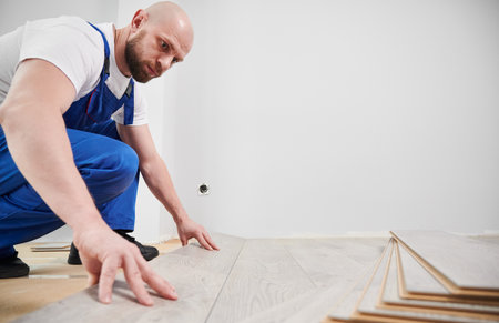 Male construction worker laying laminate timber flooring in living room with white wall on background. Man in work overalls installing laminate floor in apartment under renovation.の写真素材