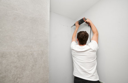 Back view of male person in white shirt using electric drilling machine while working on home renovation. Man drilling white wall with power drill tool before plumbing works in bathroom at home.の写真素材