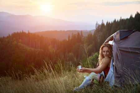 Woman tourist camping in mountains at sunset. Beautiful female sitting in tents entrance on grass with cup of tea. Young tourist hiking, having rest in campsite. Concept of tourism and hiking.の写真素材