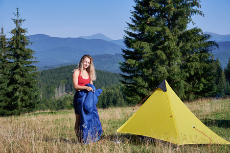 Woman tourist camping in mountains. Sporty woman hiking in summer morning outdoors. Slim, young female standing near tent, holding sleeping bag, putting in backpack. Concept of tourism and adventureの写真素材