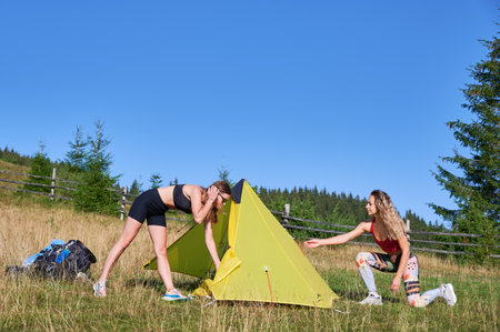 Two women hikers camping outdoors. Two female tourists hiking together, traveling in mountains. Friends wearing sportswear, putting tent, making campsite. Concept of tourism and hiking.の写真素材