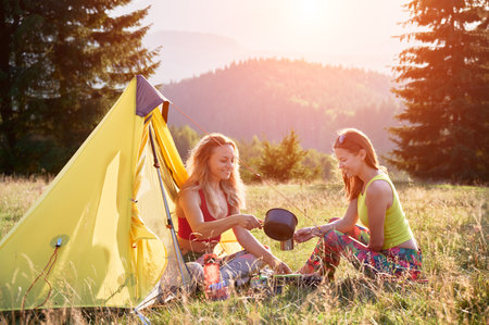 Two women tourists camping outdoors. Friends traveling, hiking outdoors in summer. Young females sitting on grass near tent in campsite, making tea, pouring water. Concept of beauty of nature.の写真素材