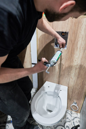Man plumber using plumbing tool applicator gun while installing toilet flush system in apartment under renovation. Male worker applying silicone sealant on corners of toilet flush panel.の写真素材