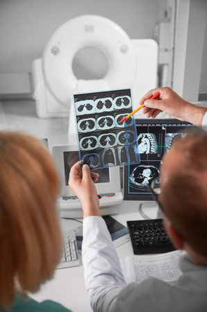 Medical computed tomography or MRI scanner. Back view of doctor and nurse sitting at computer, looking at MRI results. Doctor holding scan, showing, pointing by pen, studying, examining.の写真素材