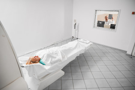 Medical computed tomography or MRI scanner. Doctor examining MRI results, female patient lying on couch. Doctor wearing glasses, holding MRI results, studying, thinking. Concept of modern diagnostics.の写真素材