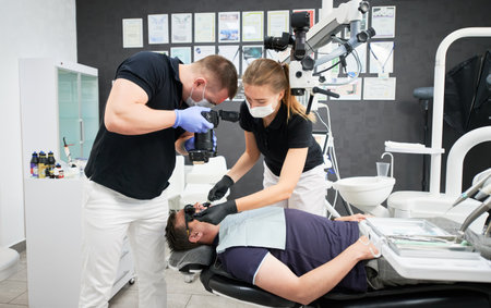 Side view of dentists fixing teeth, taking photo of result. Doctors wearing black and white uniform, masks and gloves, working in dental clinic. Concept of healthcare, dentistry.の写真素材