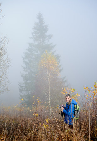 Male traveler with professional camera standing in grassy field with misty trees and fog on backgroundの写真素材