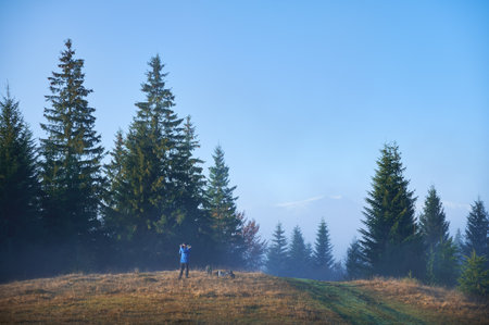 Male photographer hiker standing on grassy hill and taking picture with professional camera under blue skyの写真素材