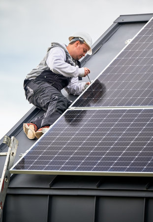 Man technician mounting photovoltaic solar panels on roof of house. Engineer in helmet installing solar module system with help of hex key. Concept of alternative, renewable energy.の写真素材