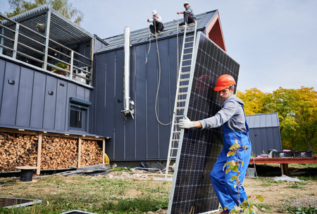 Builders building solar panel system on roof of house. Man worker in helmet carrying photovoltaic solar module outdoors. Concept of alternative and renewable energy.の写真素材