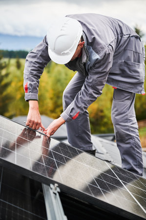 Man technician mounting photovoltaic solar panels on roof of house. Engineer in helmet installing solar module system with help of hex key. Concept of alternative, renewable energy.の写真素材