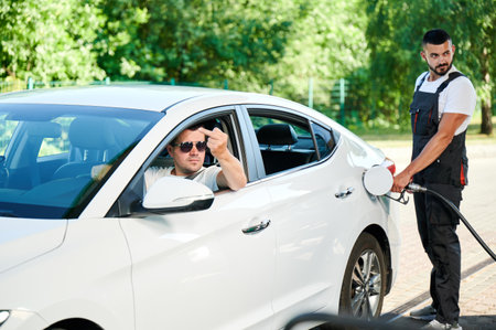 Rude driver showing middle finger to other car on gas station. Angry man in expensive car showing an indecent gesture through window. Gas station operator smiling at rude client.の写真素材
