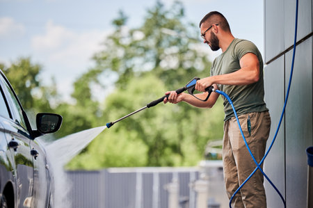 Man cleaning his car at self-service car wash with high pressure water jet. Confident man washing his blue auto with water gun at sunny day.の写真素材