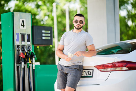 Young man waiting for petrol station operator to pay for fuel. Man leaning on his car and counting how much gasoline costs. Male in casual clothes holding money and looking straight into camera.の写真素材