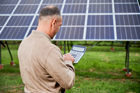 Professional investor calculating his possible profit from alternative enegry sources. Man using calculator to count exact sum of income. Back view of man counting on background of solar panels.の写真素材