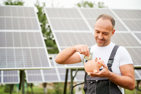 Male adult making investment in future. Knowledgeable man investing in alternative energy sources. Young man putting money in piggy bank on background of solar panels.の写真素材