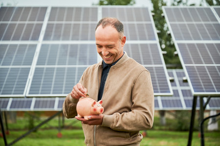 Male putting his money in piggy bank for future profit. Young man making investment in alternative ecological resources. Man in casual clothes holding moneybox on background of solar panel.の写真素材
