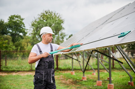 Male adult carefully wiping dust from PV solar panel. Professional worker taking care about battery cleanliness. Worker in helmet and overalls cleaning solar batteries.の写真素材