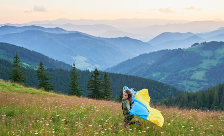 Happy Ukrainian running in Carpathian mountains. Little kid smiling and flattering Ukrainian flag on background of hills at sunset.の写真素材