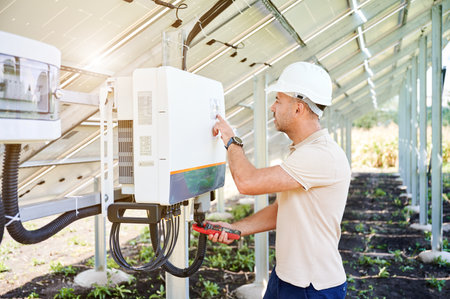 Professional worker checking voltage on solar inverter. Male using current probe to measure output voltage. Man in helmet pointing at inveter dial on back side of PV panel.の写真素材
