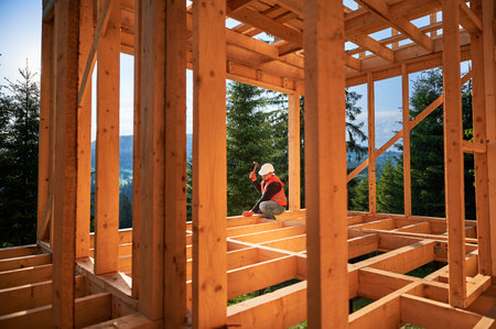 Carpenter constructing two-story wooden frame house near forest. Man hammering nails into the structure, wearing protective helmet and construction vest. Concept of modern ecological construction.の写真素材
