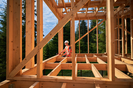 Carpenter constructing wooden frame two-story house near the forest. Man in glasses holding hammer, dressed in protective helmet and orange safety vest. Concept of ecological modern construction.の写真素材
