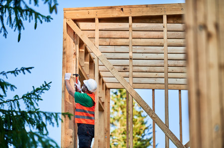 Carpenter constructing wooden frame house near the forest. Bearded man hammering nails into structure while wearing protective helmet and construction vest. Concept of modern ecological construction.の写真素材