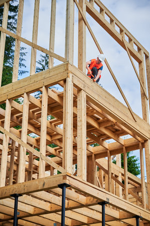 Wooden two-story frame house being built by carpenter. Man wearing glasses hammering nails using hammer, dressed in protective helmet and construction vest. Concept of modern ecological construction.の写真素材