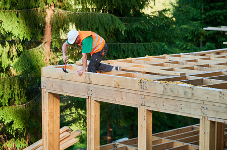 Carpenter constructing wooden frame house near the forest. Man hammering nails into structure while wearing protective helmet and construction vest. Concept of modern ecological construction.の写真素材