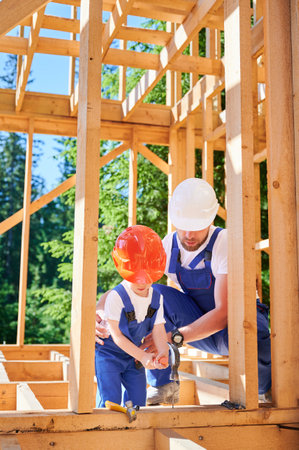 Father with toddler son building wooden frame house. Male worker tutoring his son on method of hammering nails into place, wearing helmets and blue overalls on sunny day. Carpentry and family conceptの写真素材