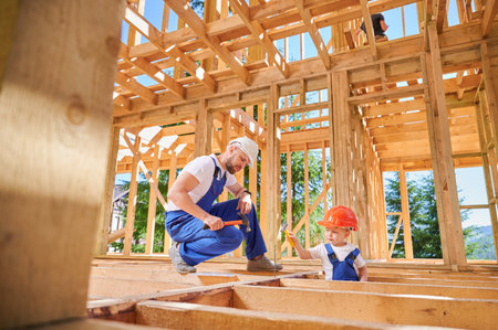 Father with toddler son constructing wooden frame house. Man builder instructing his son on art of hammering nails on construction site, wearing helmets and blue overalls. Carpentry and family conceptの写真素材