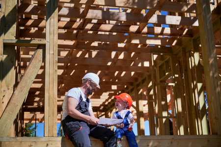 Father with toddler son building wooden frame house. Boys having fun on the edge of balcony, examining the construction plan, wearing helmets and overalls on sunny day. Carpentry and family concept.の写真素材
