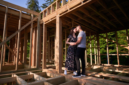 Man and woman inspecting their future wooden frame house nestled near the forest. Youthful couple at construction site in early morning. Concept of contemporary ecological construction.の写真素材